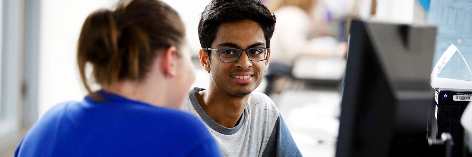 A student looks at another student and smiles while he works at a computer station.